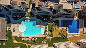 Ein Luftbild eines Hotels mit mehreren Schwimmbecken, darunter ein zentral gelegenes, kreisförmiges Becken, umgeben von viel Grün.