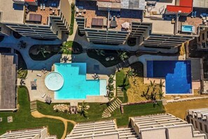 Ein Luftbild eines Hotels mit mehreren Schwimmbecken, darunter ein zentral gelegenes, kreisförmiges Becken, umgeben von viel Grün.