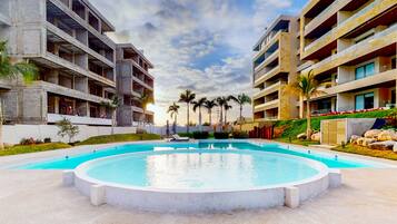 A modern hotel with a central swimming pool, multiple balconies, and palm trees.