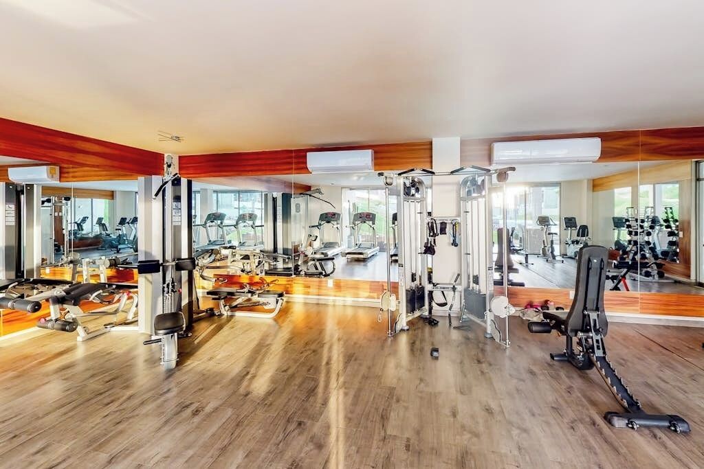 A gym with various exercise machines, wooden flooring, and large windows allowing natural light.