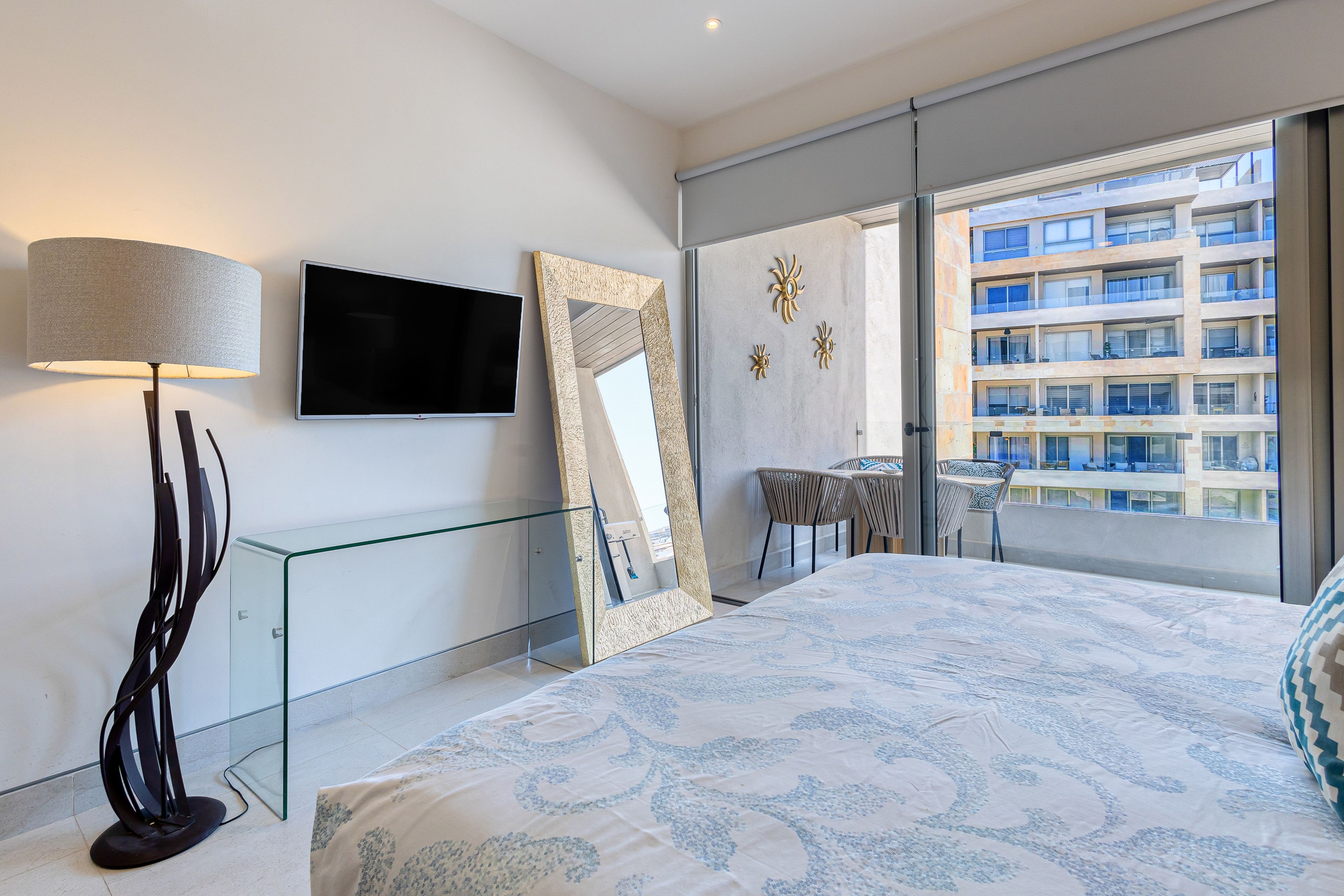 A modern bedroom with a large bed, a glass desk, a TV, and a balcony with a view of a building.