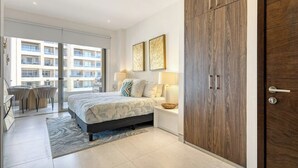 Ein Schlafzimmer mit Bett, Nachttischlampen, einem Holzschrank, einem Balkon mit Blick auf andere Gebäude sowie einem Essbereich mit Stühlen und Tisch.