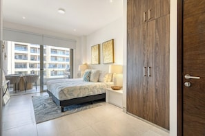 Ein Schlafzimmer mit Bett, Nachttischlampen, einem Holzschrank, einem Balkon mit Blick auf andere Gebäude sowie einem Essbereich mit Stühlen und Tisch.