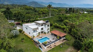 Una casa moderna de dos pisos con piscina, rodeada de vegetación exuberante y montañas al fondo.