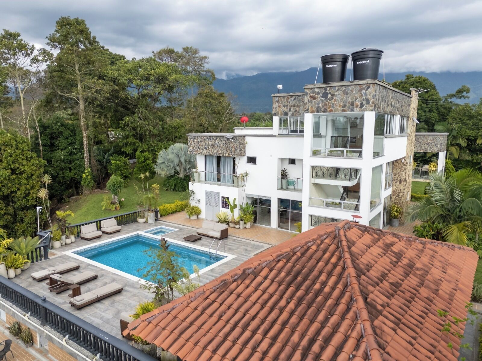A modern two-story building with a swimming pool, stone facade, and a view of lush greenery and mountains.