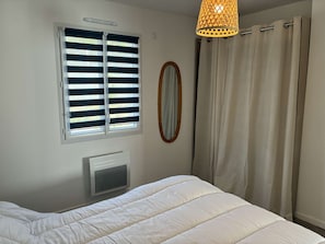 Ein Schlafzimmer mit Bett, einem Fenster mit Jalousien, einem Heizkörper, einem Spiegel und einer Hängelampe.