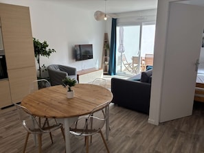 Ein modernes Wohnzimmer mit einem Holzesstisch, klaren Stühlen, einem grauen Sofa, einem an der Wand befestigten Fernseher und Blick ins Freie durch eine Glastür.