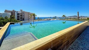 A rectangular infinity pool with clear blue water, surrounded by a tiled deck, and overlooking a coastal landscape with buildings and palm trees. - Stunning Oceanfront Luxury Apartment With Spacious Balcony Perfect for Families (Cabo San Lucas)