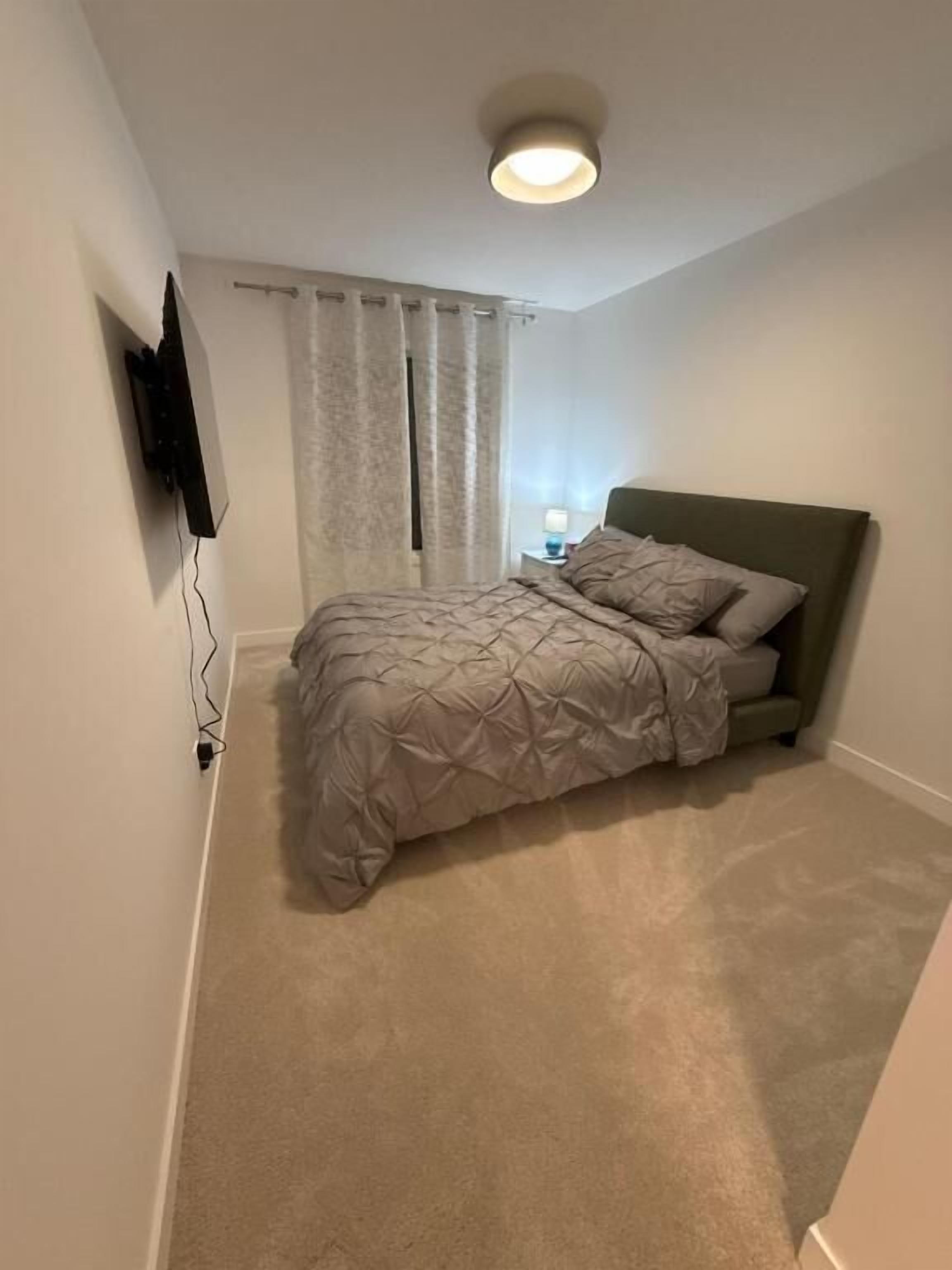 A bedroom with a bed, a nightstand, a lamp, a TV mounted on the wall, and a ceiling light.