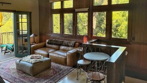 A cozy room with a leather sofa, a small round table, and a chair. There are large windows with a view of trees. - Charming furnished "Tree House" in the Hollywood Hills with views and parking. (Los Angeles)