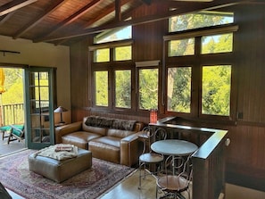 A cozy room with a leather sofa, a small round table, and a chair. There are large windows with a view of trees.