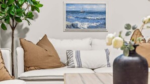 Uma sala de estar com sofá, almofadas, um vaso com flores, uma pintura do mar e uma planta.