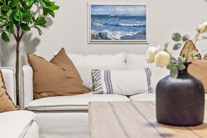 A living room with a sofa, pillows, a vase with flowers, a painting of the sea, and a plant. - The Royal Bay Stay,  by Victoria Prime! (Victoria)