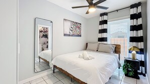 Una camera da letto con un letto, un ventilatore a soffitto, uno specchio, un quadro, una finestra con tende e un comodino con una lampada.