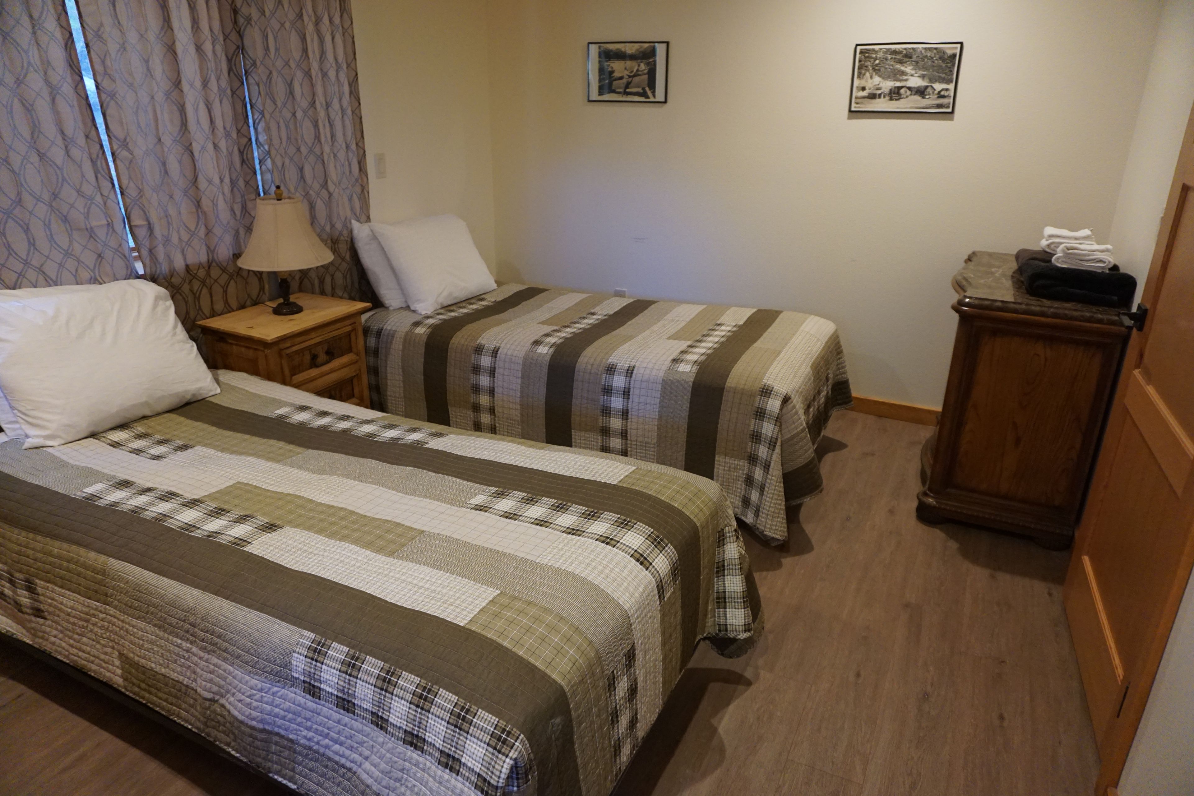 A hotel room with two single beds, a nightstand, a lamp, a small table with towels, and two framed pictures on the wall.