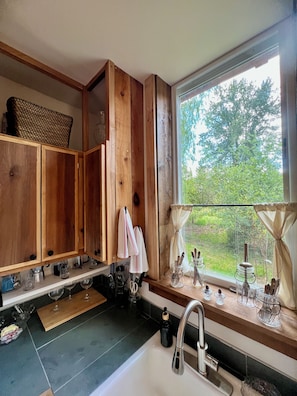 A kitchen with wooden cabinets, a window with a view of greenery, and a sink with utensils.