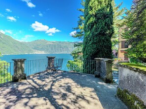 A lakeside terrace with a stone balustrade, a table with chairs, and a view of a mountain and lake. - Casa Ambra by Interhome (Nesso)
