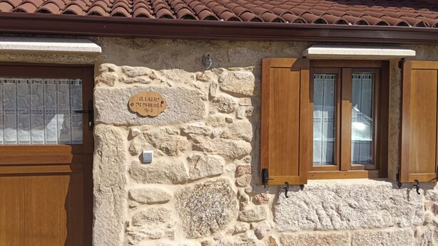 A stone building with a wooden door and window shutters, a sign on the wall, and a tiled roof.