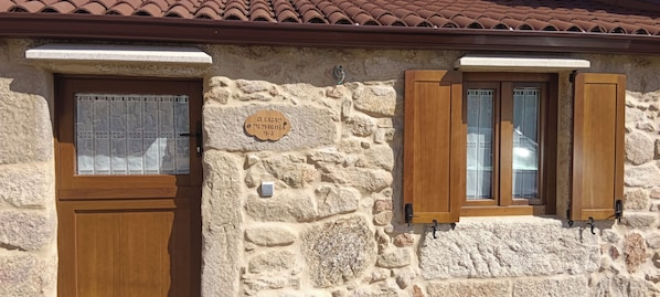 Un bâtiment en pierre avec une porte et des volets en bois, une enseigne sur le mur et un toit recouvert de tuiles.