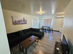 A living room with a sectional sofa, a coffee table, a painting, a dining area with a table and chairs, and an air conditioning unit.