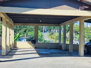 A covered outdoor area with concrete pillars, a black grill, and a view of a road and greenery.