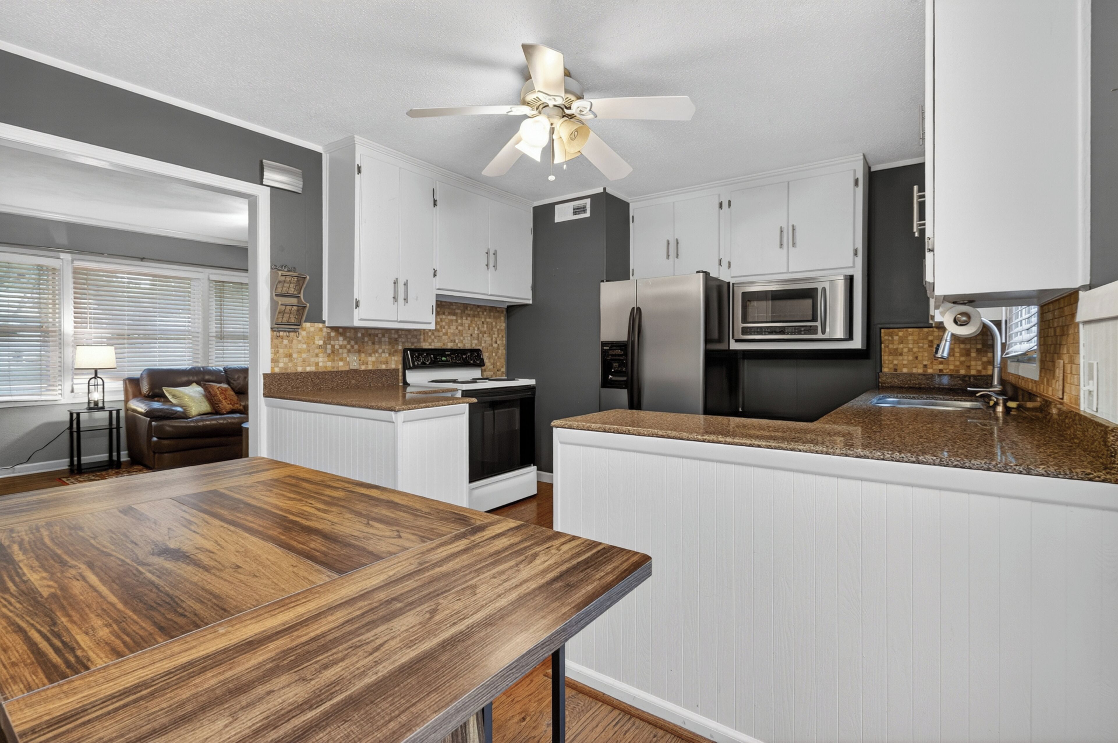 A modern kitchen with white cabinetry, a granite countertop, a built-in oven, a microwave, and a ceiling fan.