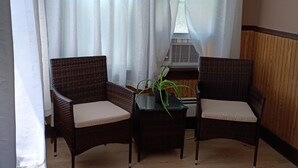 A room with two wicker chairs, a small table, a potted plant, and a window with white curtains.