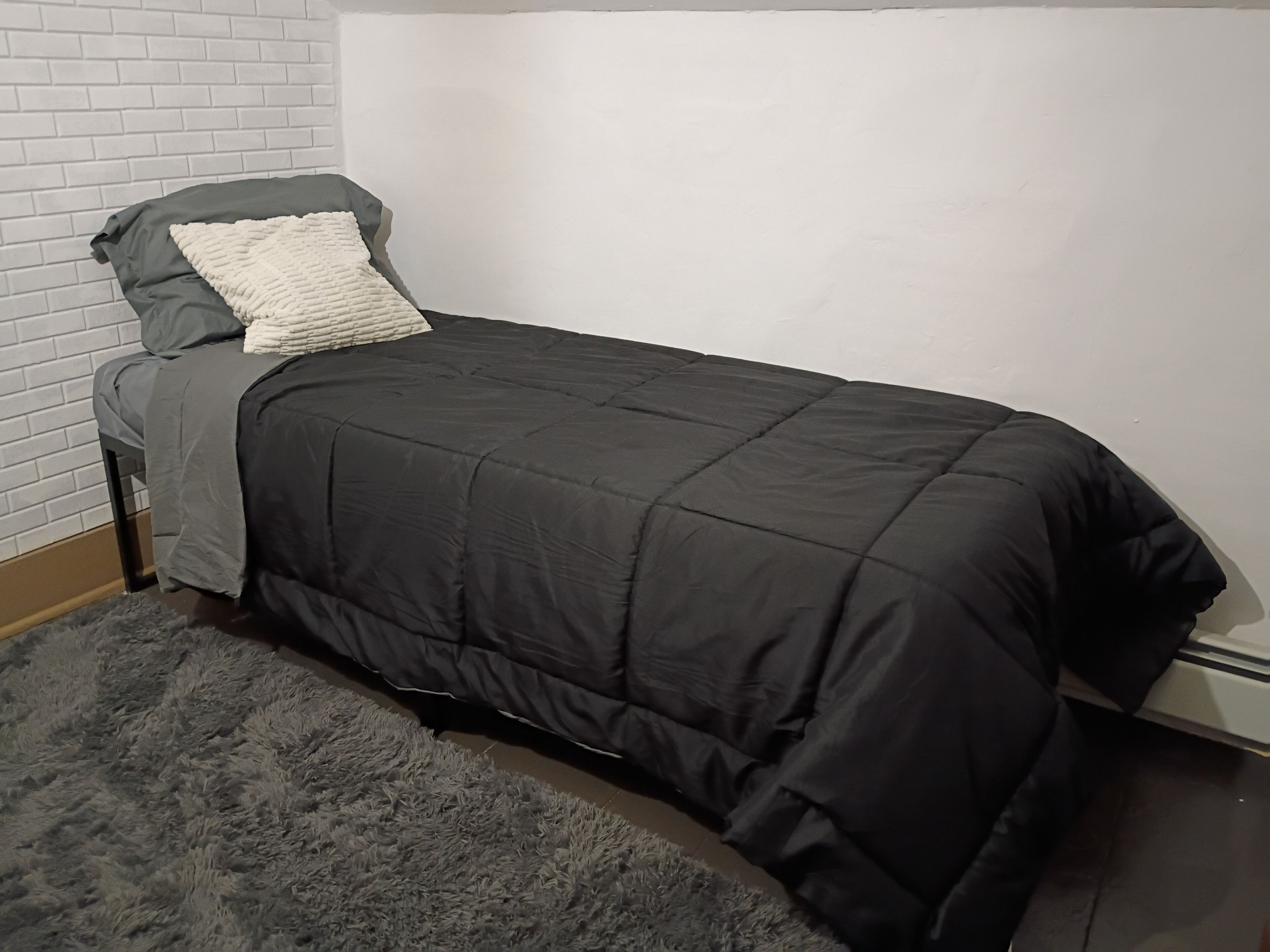 Un lit simple avec une couette et des oreillers de couleur sombre, un tapis gris à poils longs et un mur en briques blanches.