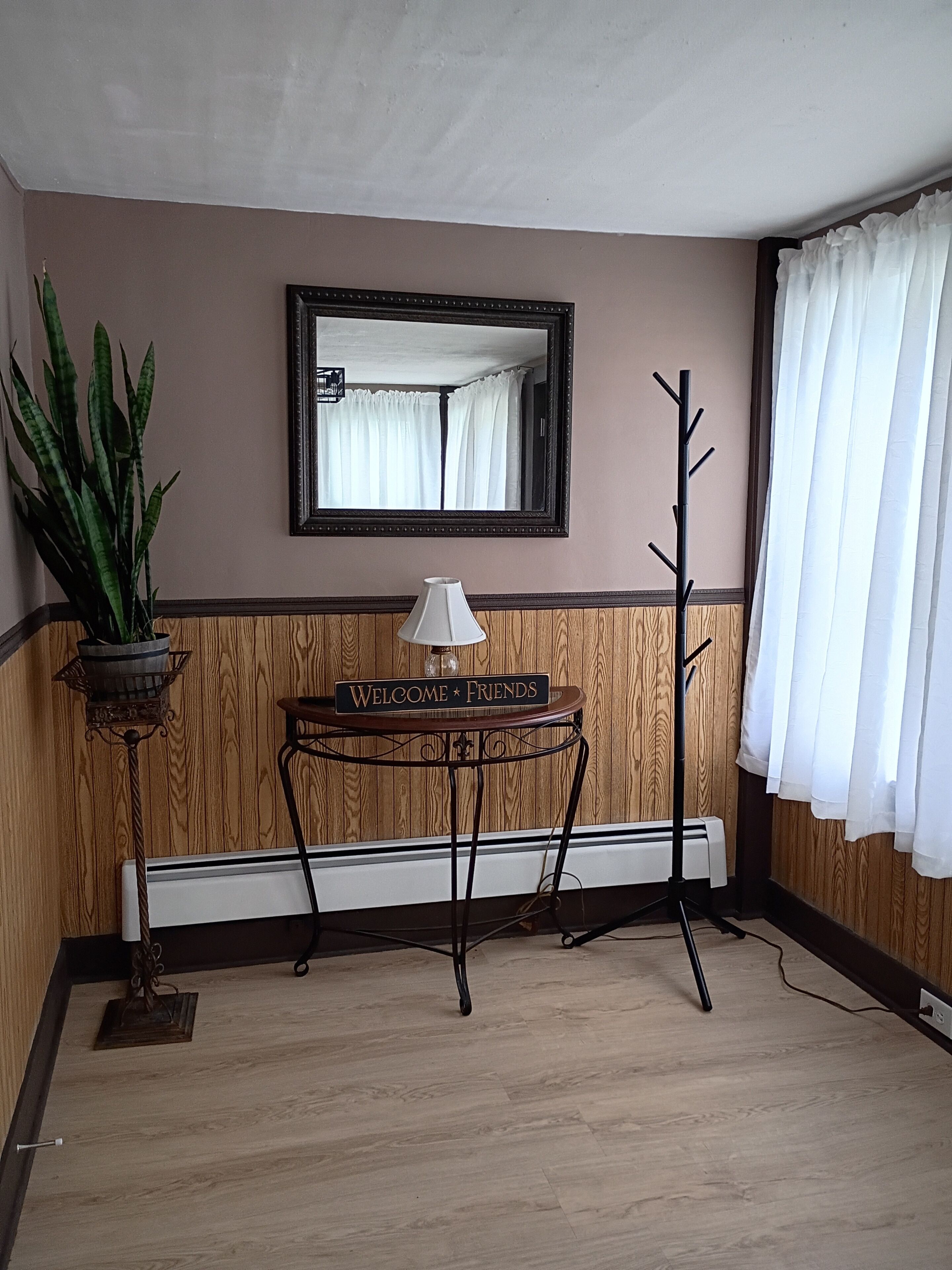 A room with a wooden floor, a radiator, a table with a lamp, a mirror, a coat rack, and a potted plant.