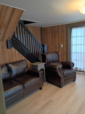 A living room with a wooden staircase, leather sofas, a small table, and a window with curtains.