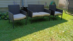 A set of wicker patio chairs with cushions, positioned on a grassy lawn in front of a house with a window and a chain-link fence.