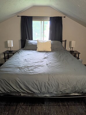 A single bed with a grey duvet, two bedside tables with lamps, a window with curtains, and a sloped ceiling.