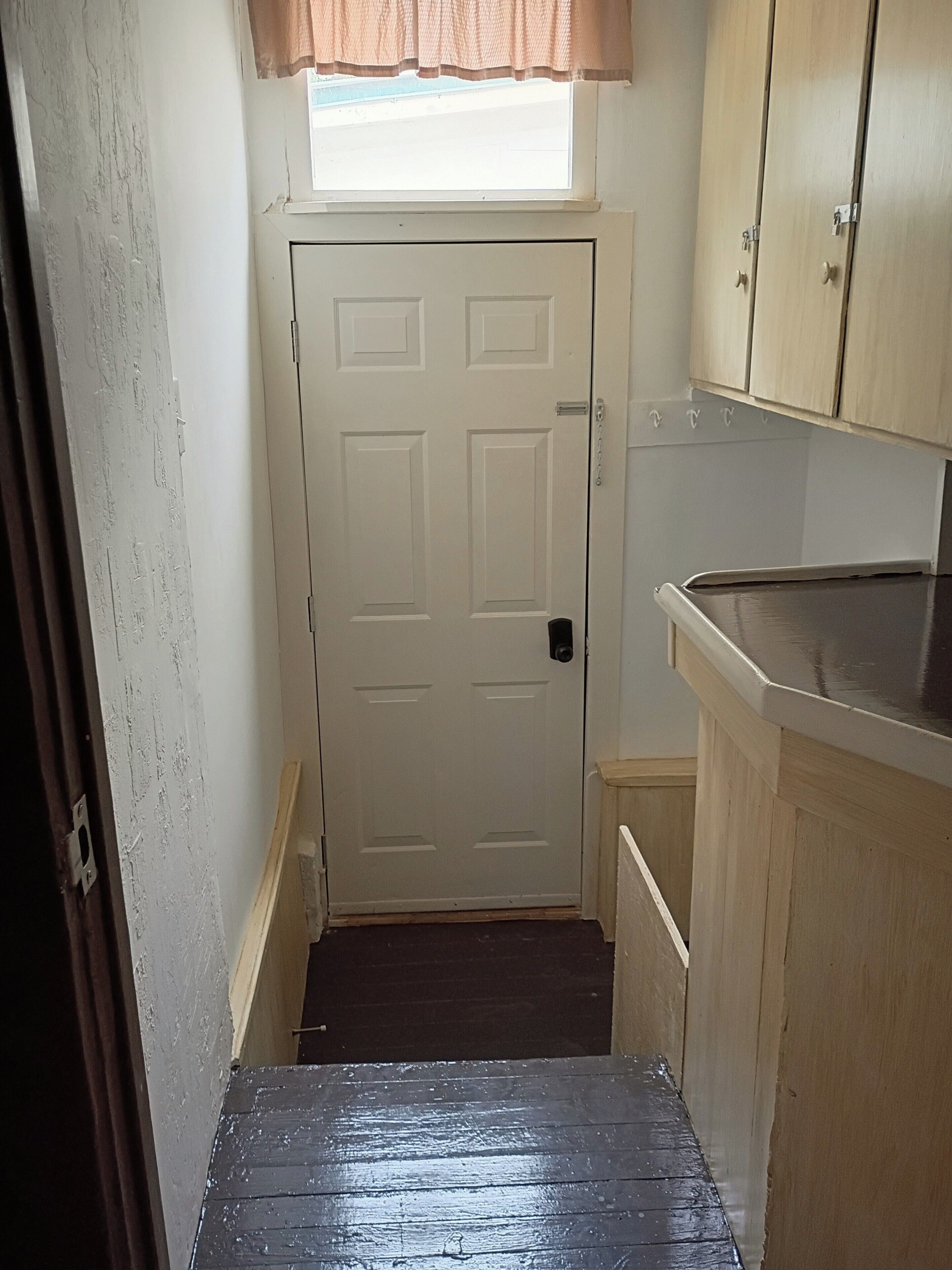 A narrow hallway with a white door, a window with a brown valance, and wooden cabinets.
