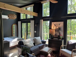Ein modernes Schlafzimmer mit Bett, Sofa und Sessel, großen Fenstern und einem Gemälde eines Pferdes.