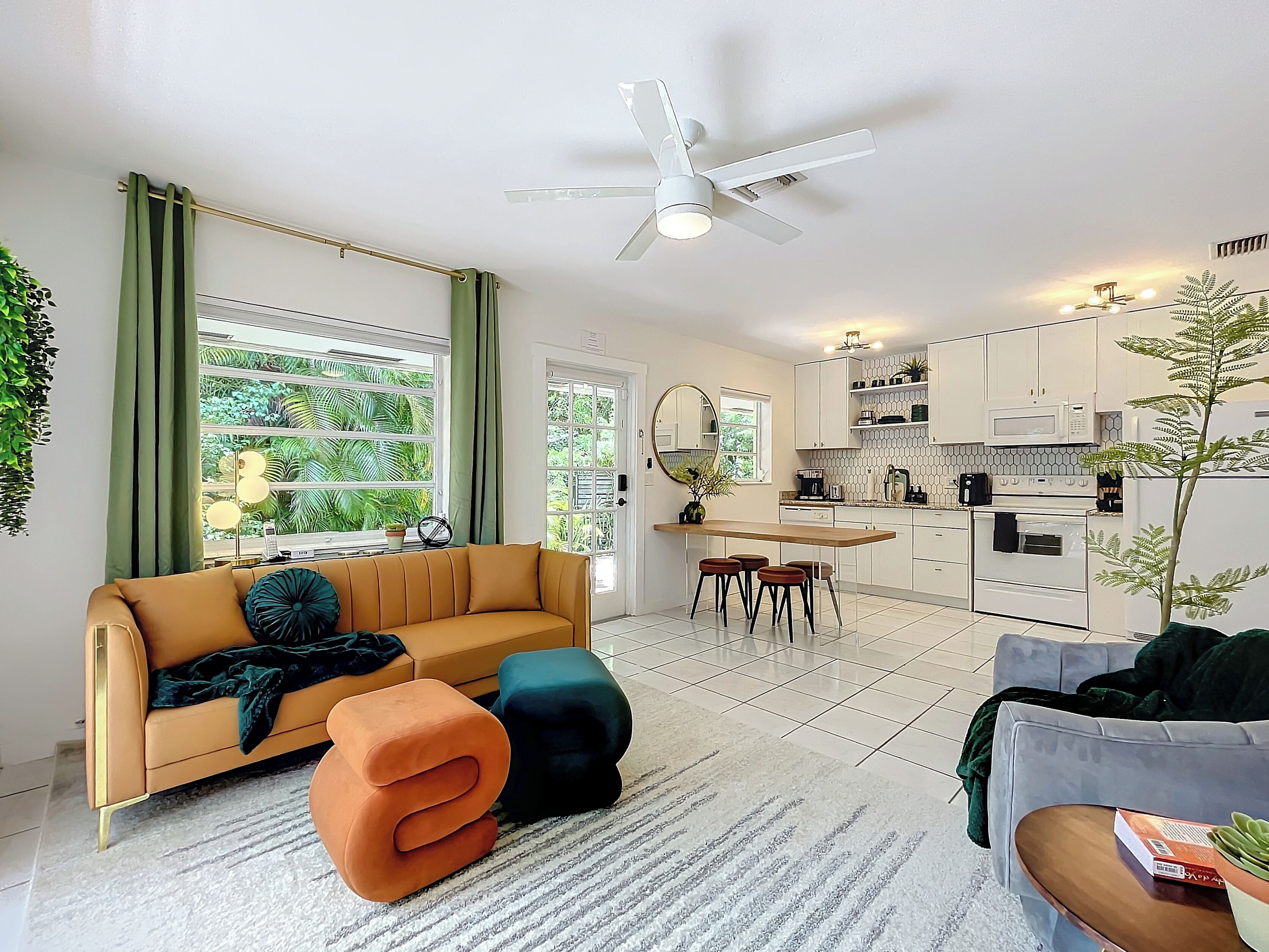 A modern living room with a sofa, ottoman, and coffee table, a dining area with a table and chairs, and a kitchen with white cabinets and appliances.