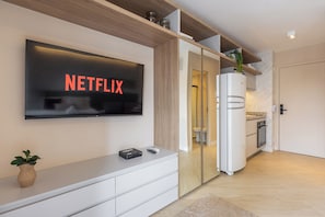 Uma sala de estar moderna com uma TV de tela plana exibindo 'NETFLIX', um móvel de entretenimento branco, uma pequena planta e uma área de cozinha com geladeira e armários.