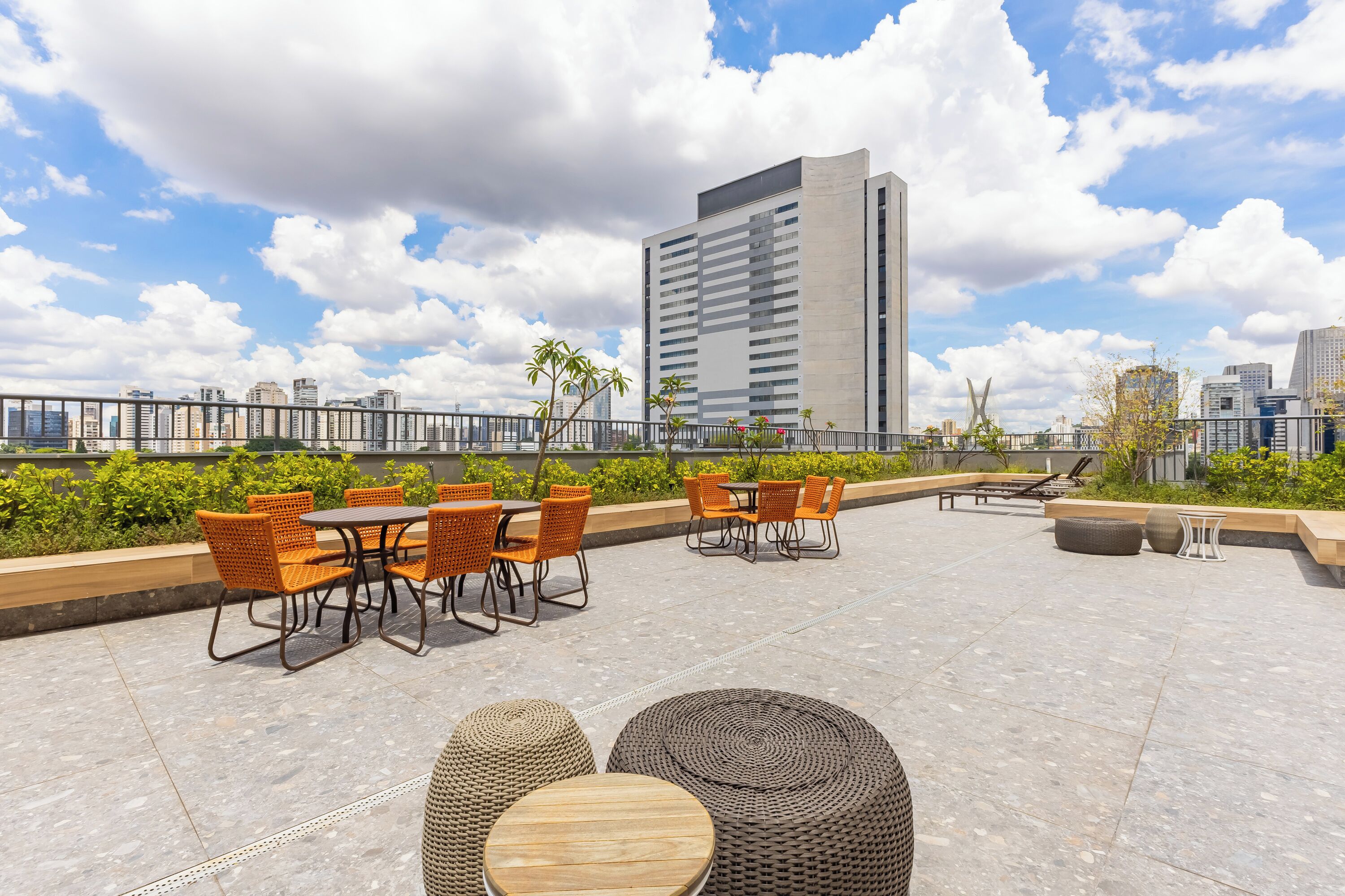 Uma área de estar externa com mesas e cadeiras, vista para o horizonte da cidade e um edifício moderno ao fundo.