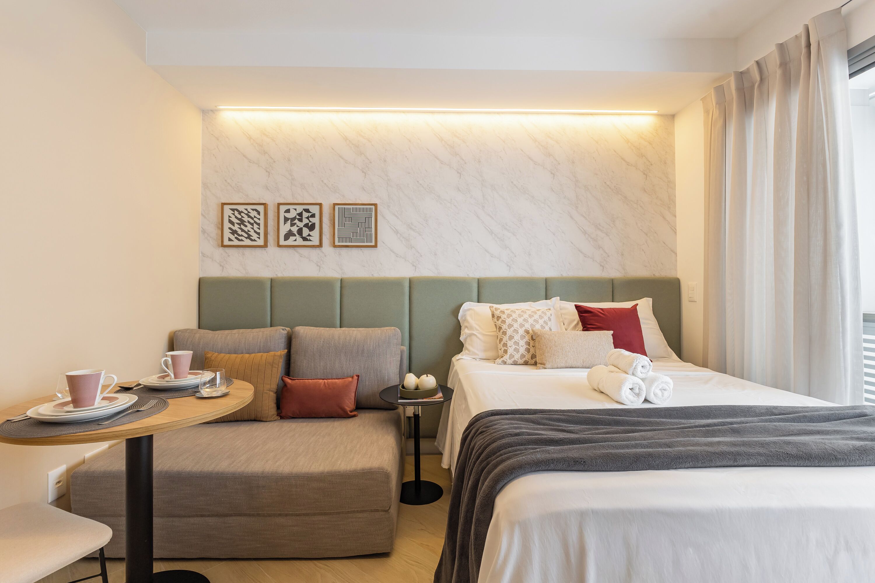 Quarto de hotel com cama, sofá, mesa pequena com xícara e pires, e parede com três quadros.