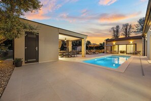 Ein moderner Poolbereich im Freien mit überdachter Sitzgelegenheit und einem Gebäude mit Naturstein-Fassade.