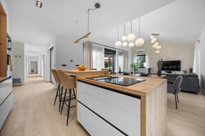 Eine moderne Küche mit einer zentralen Kochinsel, Barhocker, Pendelleuchten und einem Essbereich mit einer Couch und Fernseher.