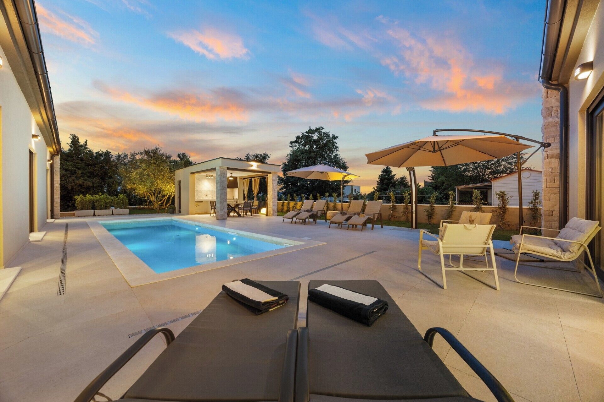 A swimming pool with lounge chairs and umbrellas, a covered seating area, and a small building with a grill.