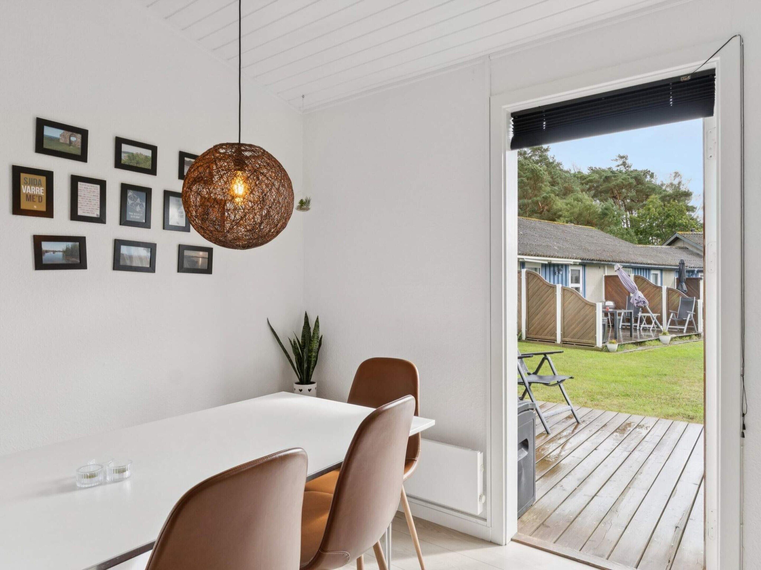 A dining area with a white table, two chairs, a hanging light fixture, and a view of a backyard with a wooden deck and outdoor furniture.