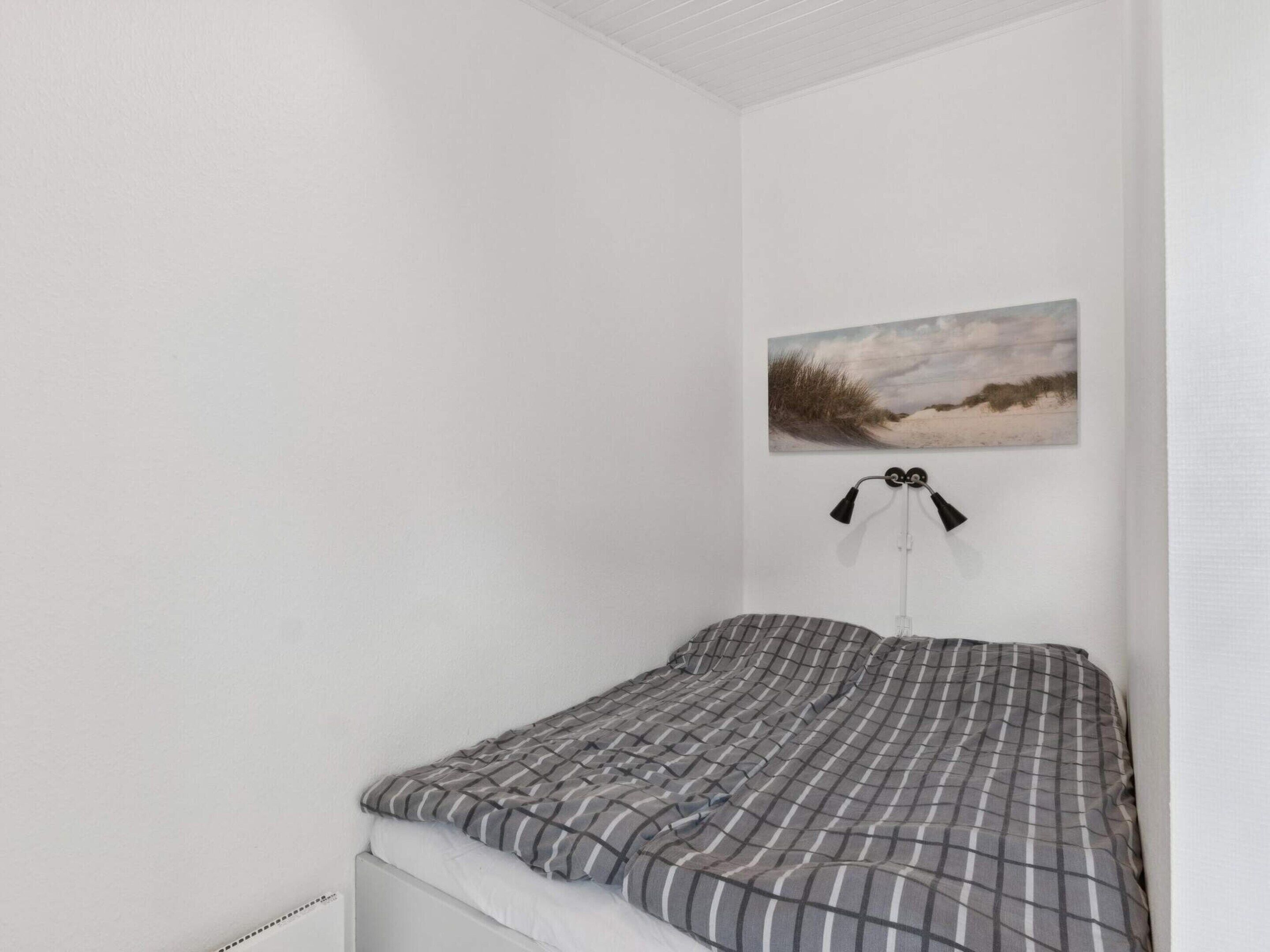 A single bed with a checkered blanket, a wall-mounted lamp, and a beach landscape painting.