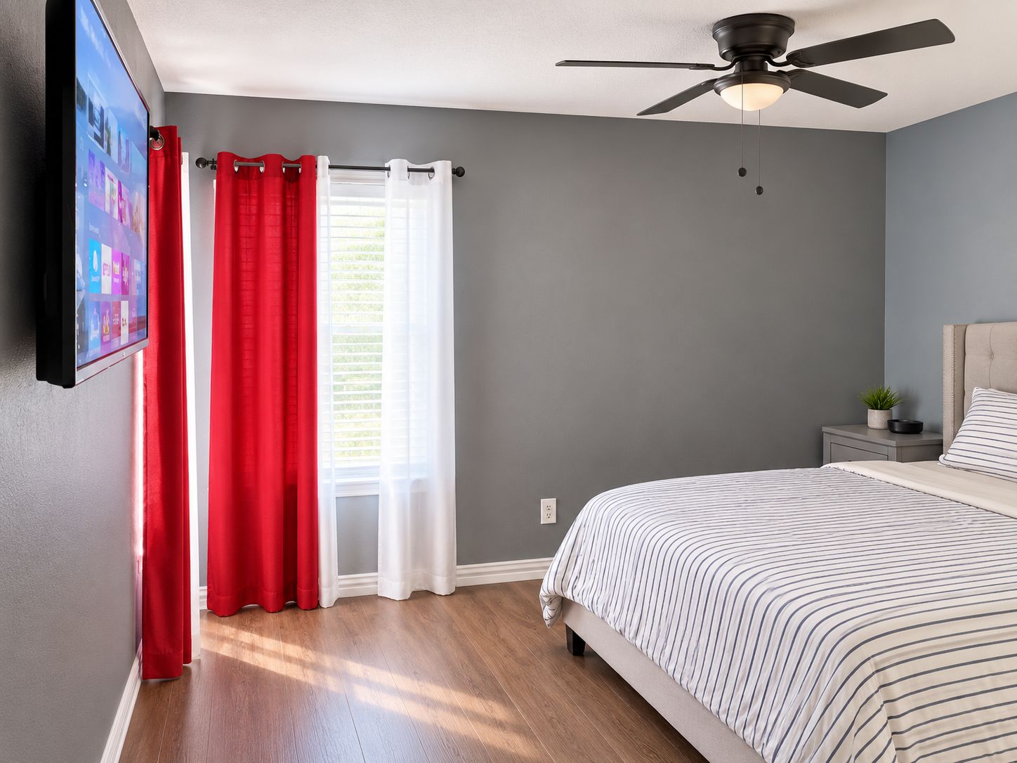 A bedroom with a bed, a ceiling fan, a nightstand with a plant, and a window with red and white curtains.