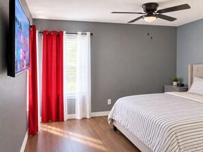 Een slaapkamer met een bed, een plafondventilator, een nachtkastje met een plant en een raam met rode en witte gordijnen.