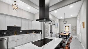 A modern kitchen with white cabinets, a black island, stainless steel appliances, and a dining area with a wooden table and chairs. - Two Dining Tables · Sleeps 14 · 7BR, 4 Baths (Fuquay-Varina)