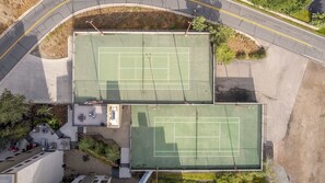Luftaufnahme eines Tennisplatzkomplexes mit zwei Plätzen, einem Parkplatz und einem Gebäude mit Dachgarten.