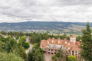 Un grand immeuble à étages, doté d’une tour imposante, entouré d’une végétation luxuriante et offrant une vue sur des collines lointaines.