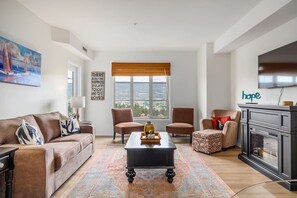 A living room with a sofa, armchairs, a coffee table, a fireplace, and a large window with a view. - 2BR | Pool, Hot Tub, Golf, UBC & Airport (Kelowna)