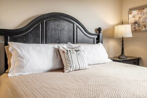 A bed with a quilted cover, two pillows, a nightstand with a lamp, and a wall with a floral painting. - 2BR | Pool, Hot Tub, Golf, UBC & Airport (Kelowna)